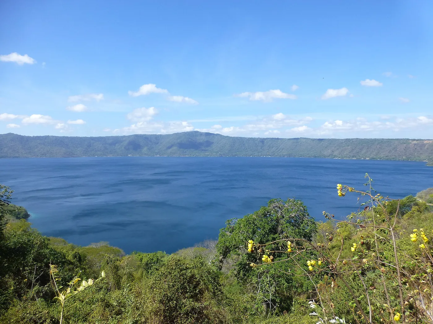A blue crater lake inside a forested caldera seen from the rim